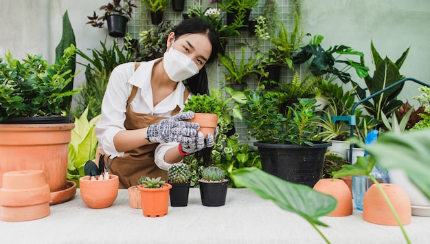 Jardinero asiático mujer con mascarilla y delantal sosteniendo una planta de interior verde en la mano mientras cuida la planta de interior de interior