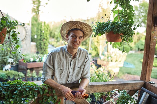 Jardinero de chico con sombrero de paja está de pie junto a una terraza de madera con muchas plantas en un día soleado. .