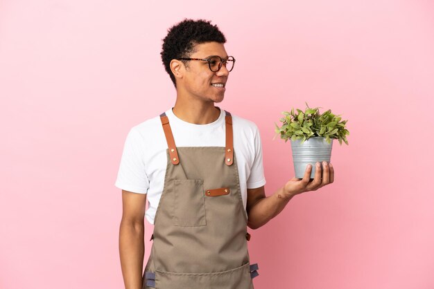 Jardinero hombre africano sosteniendo una planta aislada sobre fondo rosa mirando hacia el lado y sonriendo