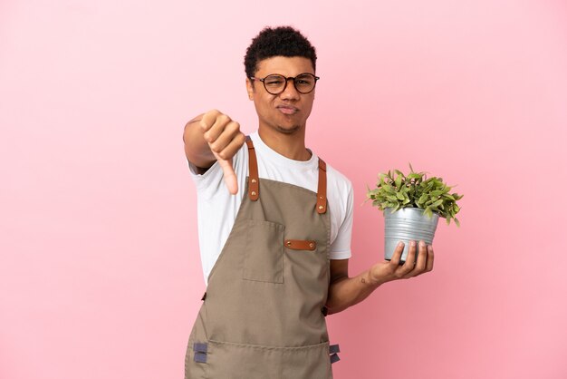 Jardinero hombre africano sosteniendo una planta aislada sobre fondo rosa mostrando el pulgar hacia abajo con expresión negativa