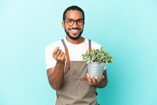 Jardinero hombre latino sosteniendo una planta aislada sobre fondo azul haciendo gesto de dinero