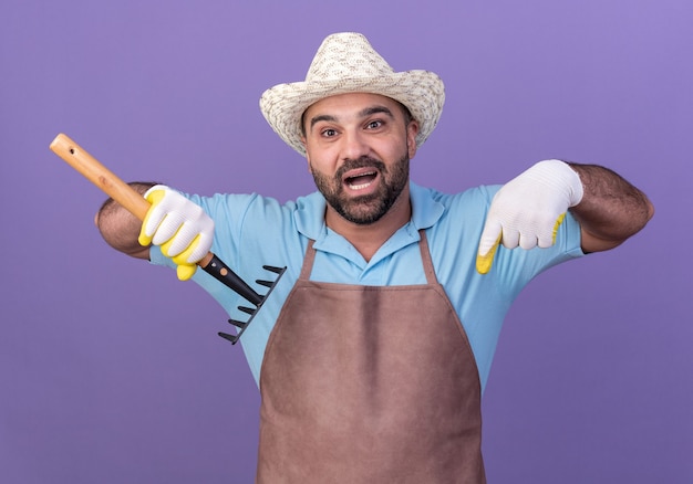 Jardinero macho caucásico adulto asustado vistiendo guantes y gorro de jardinería sosteniendo el rastrillo y apuntando hacia abajo