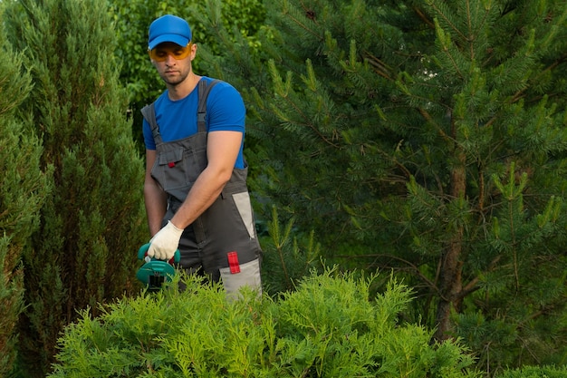 Jardinero macho en un traje de protección poda los arbustos con una podadora eléctrica