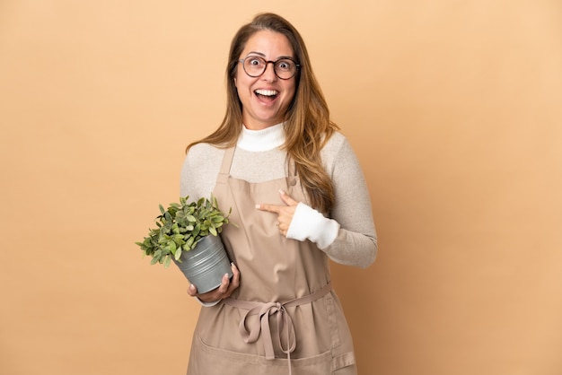 Jardinero de mediana edad mujer sosteniendo una planta aislada en pared beige con expresión facial sorpresa