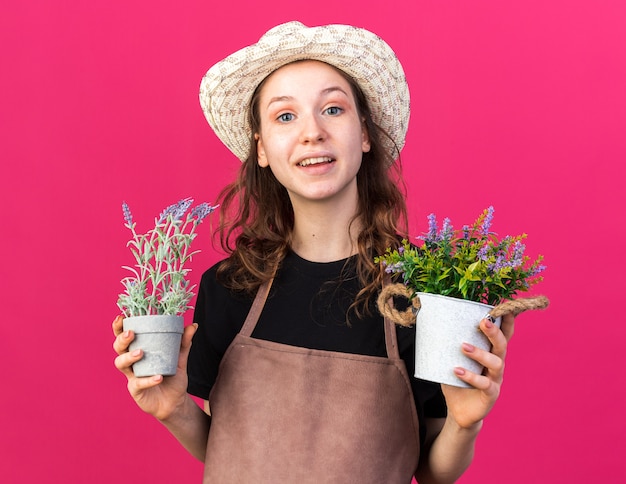 Jardinero mujer joven sonriente con sombrero de jardinería sosteniendo flores en macetas aislado en la pared rosa