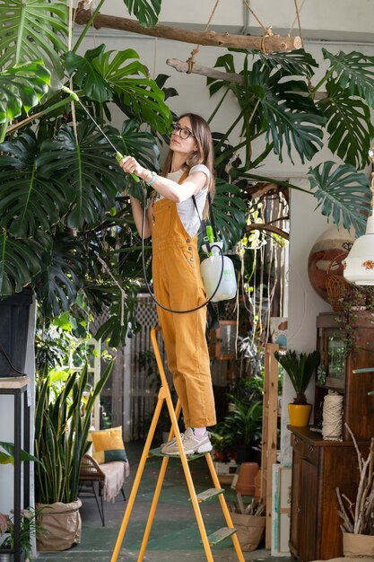 Jardinero de mujer en monos cuidando exuberante planta de monstera en el acogedor jardín de su casa