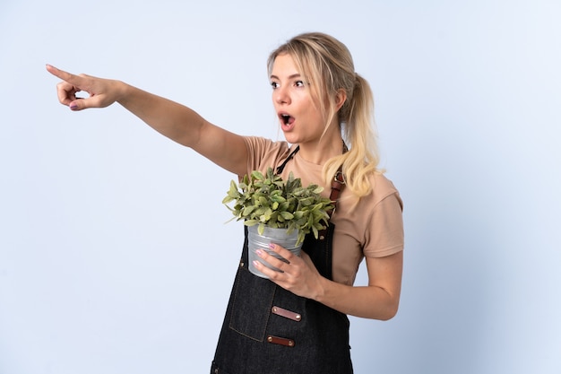 Foto jardinero rubio mujer sosteniendo una planta sobre pared aislada apuntando hacia afuera