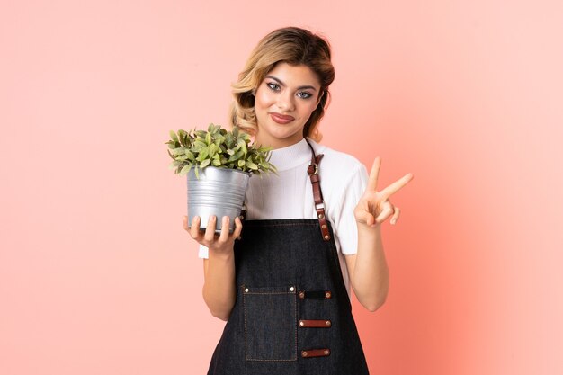 Jardinero ruso chica sosteniendo una planta aislada en rosa sonriendo y mostrando el signo de la victoria