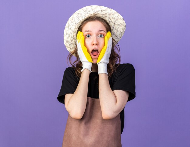 Jardinero de sexo femenino joven asustado que lleva el sombrero de jardinería con los guantes que cubren la cara con las manos aisladas en la pared azul