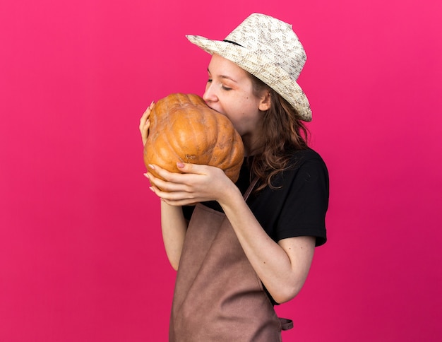 Jardinero de sexo femenino joven con sombrero de jardinería sosteniendo mordeduras de calabaza aislado en la pared rosa