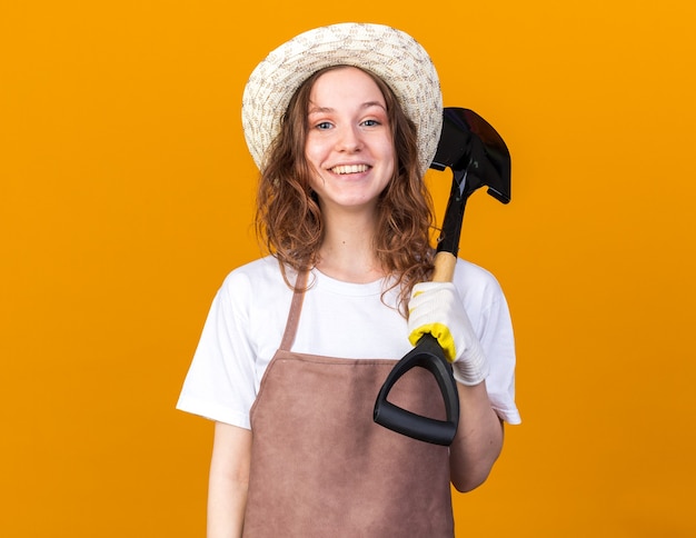 Jardinero de sexo femenino joven sonriente que lleva guantes del sombrero de jardinería que sostiene la pala en el hombro