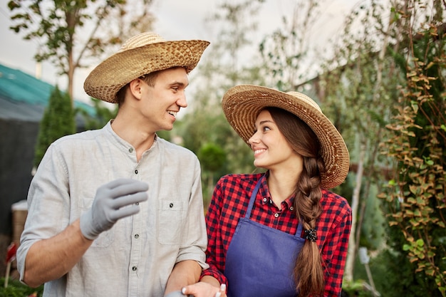 Foto jardineros de chico y chica sonrientes con sombreros de paja miran el uno al otro en el jardín en un día soleado. .