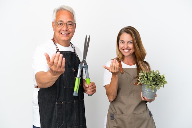 Jardineros de mediana edad sosteniendo una planta y tijeras aisladas sobre fondo blanco invitando a venir con la mano. Feliz de que hayas venido