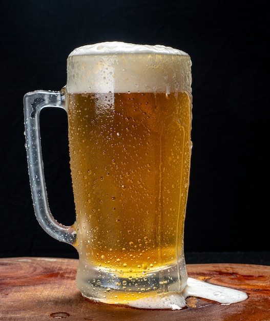 Jarra de cerveza, servida fría con abundante espuma sobre una tabla de madera sobre un fondo negro.