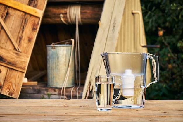 Jarra con filtro de agua y una taza de vidrio transparente con agua limpia en frente del pozo de madera al aire libre en las noches de verano