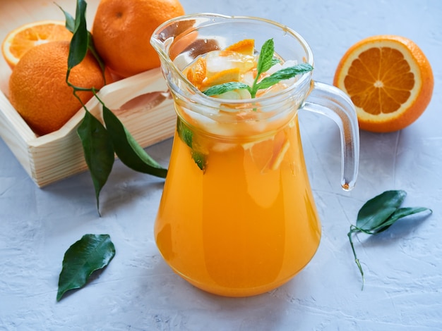 Jarra de jugo de naranja natural con rodajas de naranja, hielo, hojas de menta sobre una superficie gris