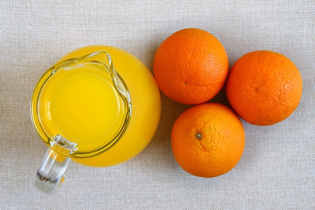 Jarra y vaso de jugo de naranja y naranjas maduras desde arriba, vista superior sobre tela de arpillera