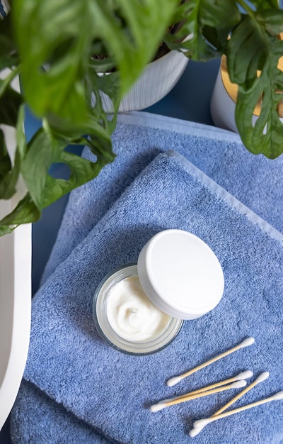 Foto jarro de crema abierto en toallas azules cerca del lavabo y monstera verde en maqueta de tapa de baño