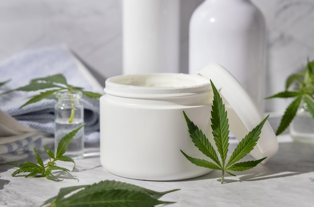 Jarro de creme branco aberto perto de folhas de cannabis verdes na casa de banho perto de outras garrafas close up mockup produtos de beleza orgânicos para a pele cosméticos naturais de CBD