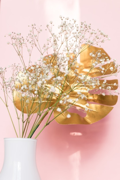 Jarrón blanco con hojas tropicales doradas y flores de gypsophila contra la pared rosa. Decoración interior con estilo