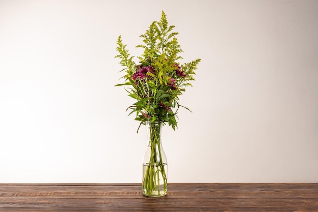 Jarrón de cristal con ramo de flores hermosas sobre mesa de madera sobre fondo de pared blanca