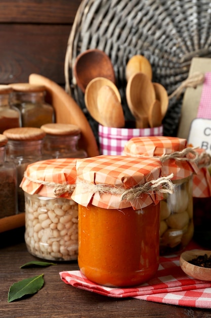 Jarros com legumes e feijões em conserva, especiarias, livros de receitas e utensílios de cozinha em fundo de madeira