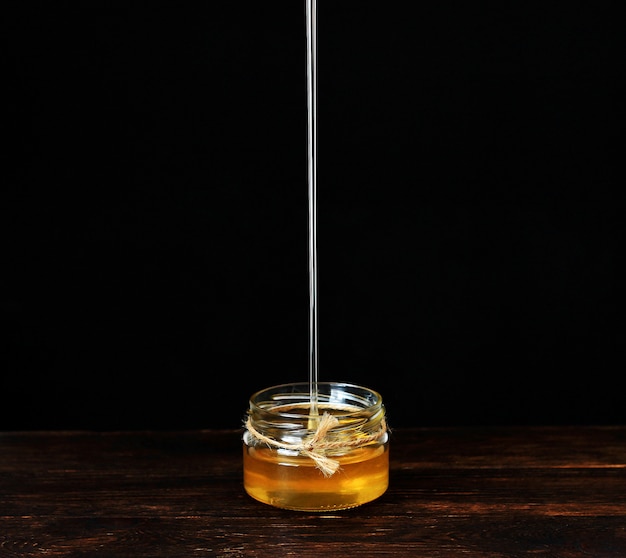 Jato de mel derramando em uma jarra de vidro, pingando mel líquido, saboroso amarelo dourado. Mesa de madeira marrom contra preto. Copyspace.