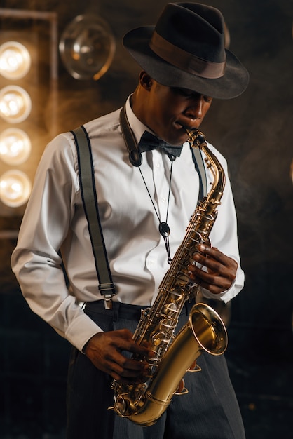 Jazzman negro con sombrero toca el saxofón en el escenario con focos. Músico de jazz negro preformando en la escena