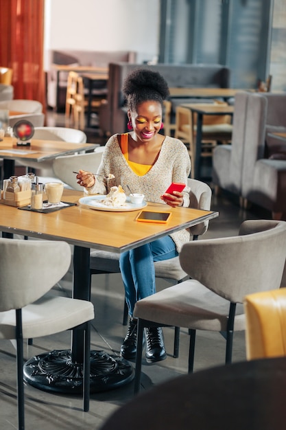 Jeans y suéter darkhaired mujer vestida con jeans y suéter ligero sentado en la cafetería comiendo pastel dulce