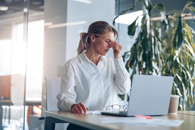 Jefa cansada con dolor de cabeza trabajando en una laptop en su lugar de trabajo en la oficina moderna