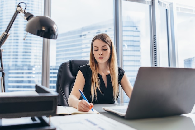 Jefa ejecutiva femenina sentada en su escritorio tomando notas en la agenda escribiendo con lápiz y usando su computadora en el edificio de oficinas moderno.