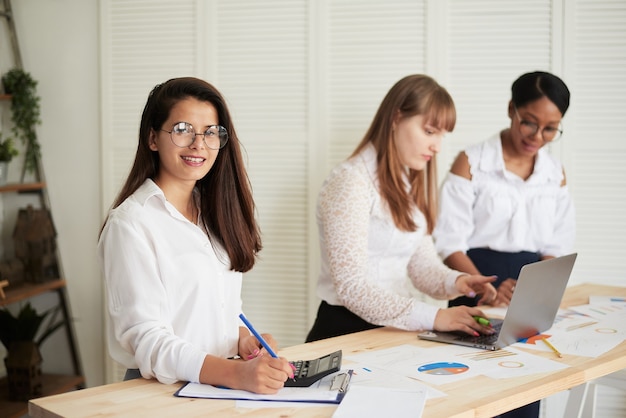 La jefa de un equipo empresarial femenino multiétnico se sienta en un escritorio, escribe en un cuaderno y mira a la cámara.