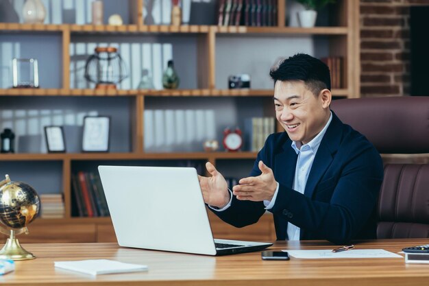 El jefe asiático exitoso y feliz mira la pantalla de la computadora portátil y sonríe en una videollamada con sus colegas para contarles los planes financieros de la reunión de negocios en línea