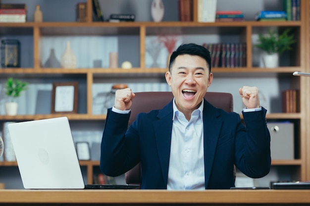 Foto el jefe asiático feliz mira la cámara y celebra la victoria empresarial levanta las manos en gesto de triunfo