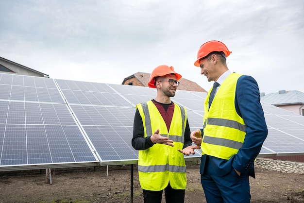 El jefe con casco y chaleco se encuentra con un empleado vestido con ropa especial cerca de los paneles solares. Concepto de electricidad verde