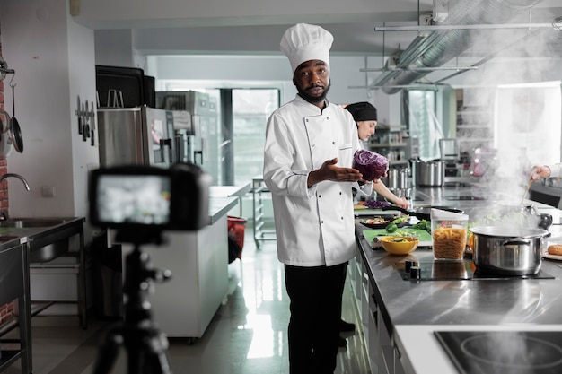 El jefe de cocina afroamericano tiene repollo rojo filmando la cocina de un plato gourmet mientras mira la cámara. Vlogueros culinarios en la cocina del restaurante grabando un curso culinario para Internet y transmisión televisiva.