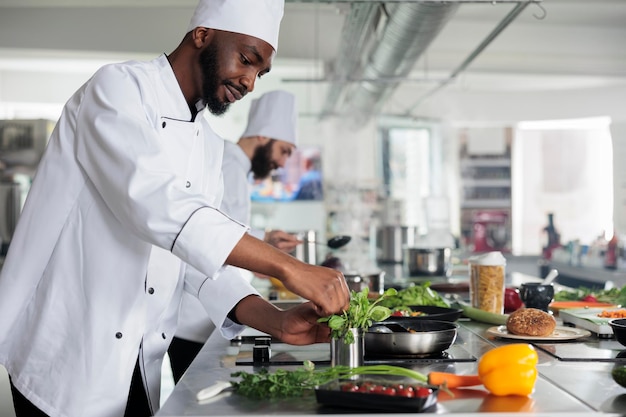 Jefe de cocina profesional afroamericano recogiendo hierbas verdes frescas para mejorar el sabor del plato gourmet. Cocinero masculino con uniforme de cocina preparando comida orgánica mientras está en la cocina del restaurante.