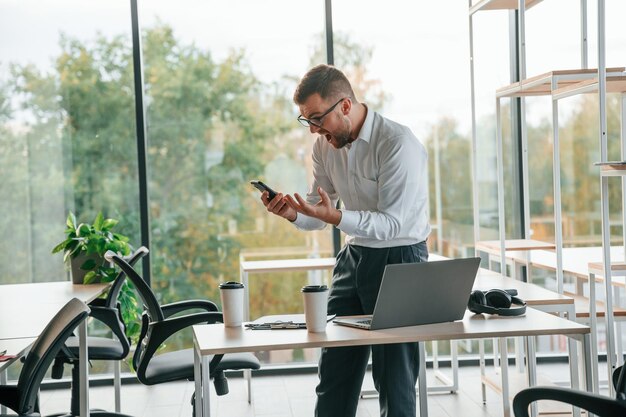 Foto el jefe enojado está de pie junto a la mesa con una computadora portátil en él. el hombre con ropa formal está trabajando.