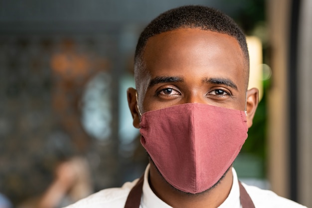 Jefe de joven camarero contemporáneo de etnia africana en máscara protectora de pie delante de la cámara contra el interior de la cafetería o restaurante