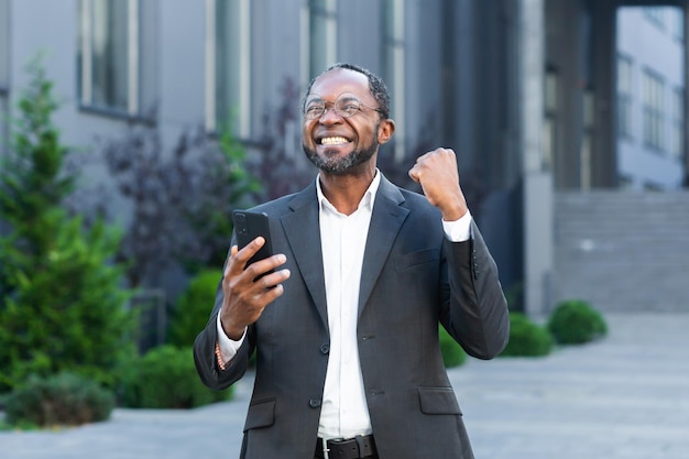 Jefe de negocios afroamericano fuera del edificio de oficinas moderno usando el teléfono senior hombre celebrando