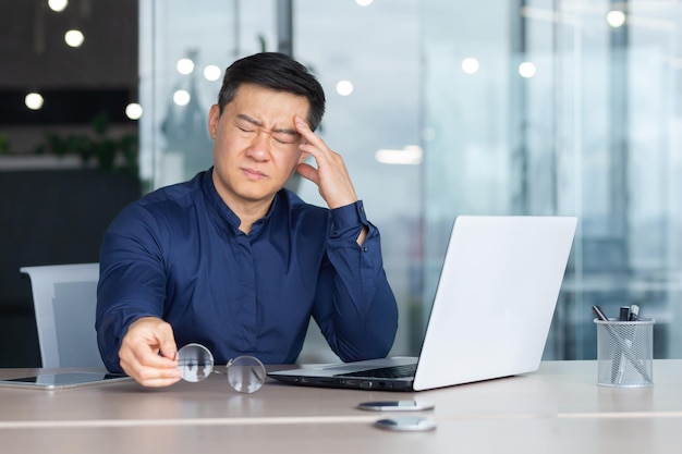 El jefe de negocios asiático enfermo tiene dolor de cabeza severo hombre con anteojos sentado en el escritorio usando una computadora portátil en el trabajo