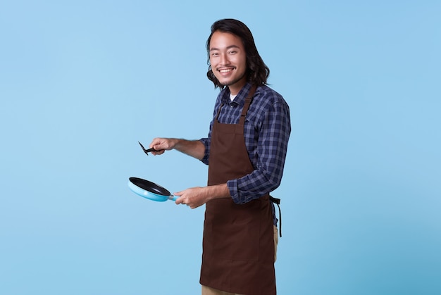 Jefe profesional asiático listo para cocinar, Chef con espátula y sartén aislado sobre fondo azul.