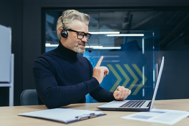 Jefe senior y experimentado de pelo gris que trabaja en un hombre de oficina moderno con auriculares hablando en video