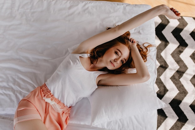 Foto jengibre aburrida acostada en una cama blanda y estirándose captura interior de una joven modelo extasiada