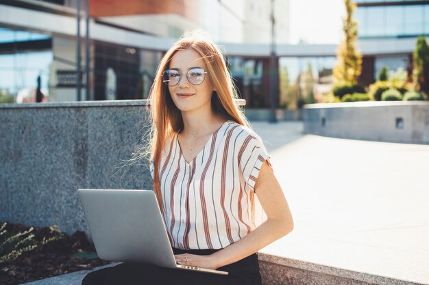 Jengibre empresaria caucásica con pecas y gafas trabajando con una computadora afuera