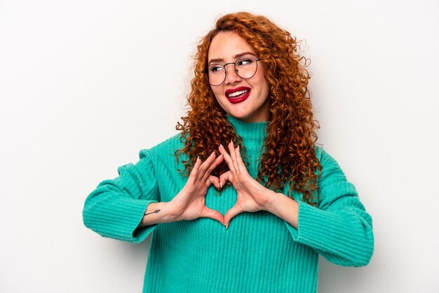 Jengibre joven mujer caucásica aislada sobre fondo blanco sonriendo y mostrando una forma de corazón con las manos