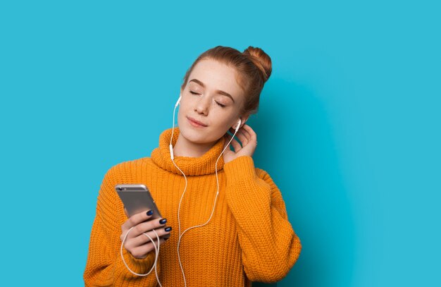 Jengibre mujer caucásica con pecas escucha alegremente meditando usando un teléfono y auriculares mientras posa en una pared azul con espacio libre