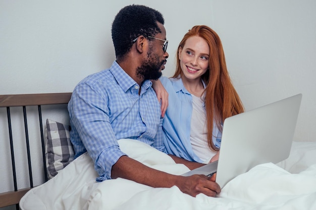 Jengibre pelirroja mujer europea y apuesto hombre afroafricano juntos abrazados acostados en el dormitorio en casa apartamento acogedor y viendo noticias en laptoplifestyle familia interracial