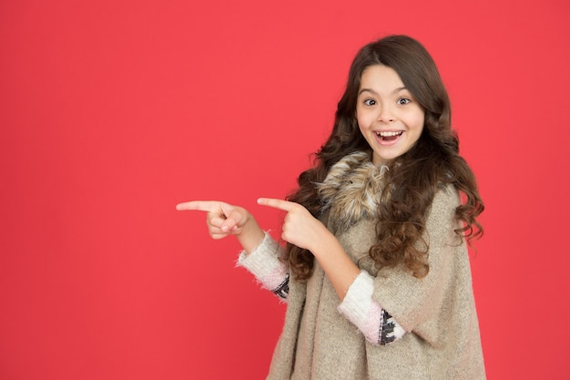 Jersey abrigado para la temporada de frio. estilo de moda infantil. infancia feliz. niño presentando producto. chica sorprendida pelo largo y rizado. pequeña belleza sonriendo sobre fondo rojo. concepto de peluquería. Copie el espacio.