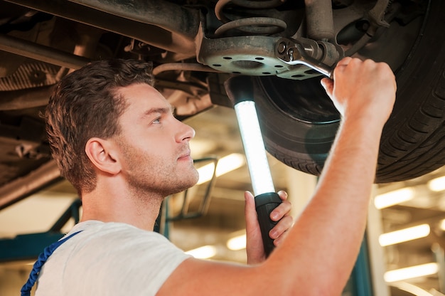 Jetzt kenne ich das Problem. Niedriger Blickwinkel eines selbstbewussten jungen Mannes in einheitlicher Haltelampe, während er in der Werkstatt unter einem Auto steht